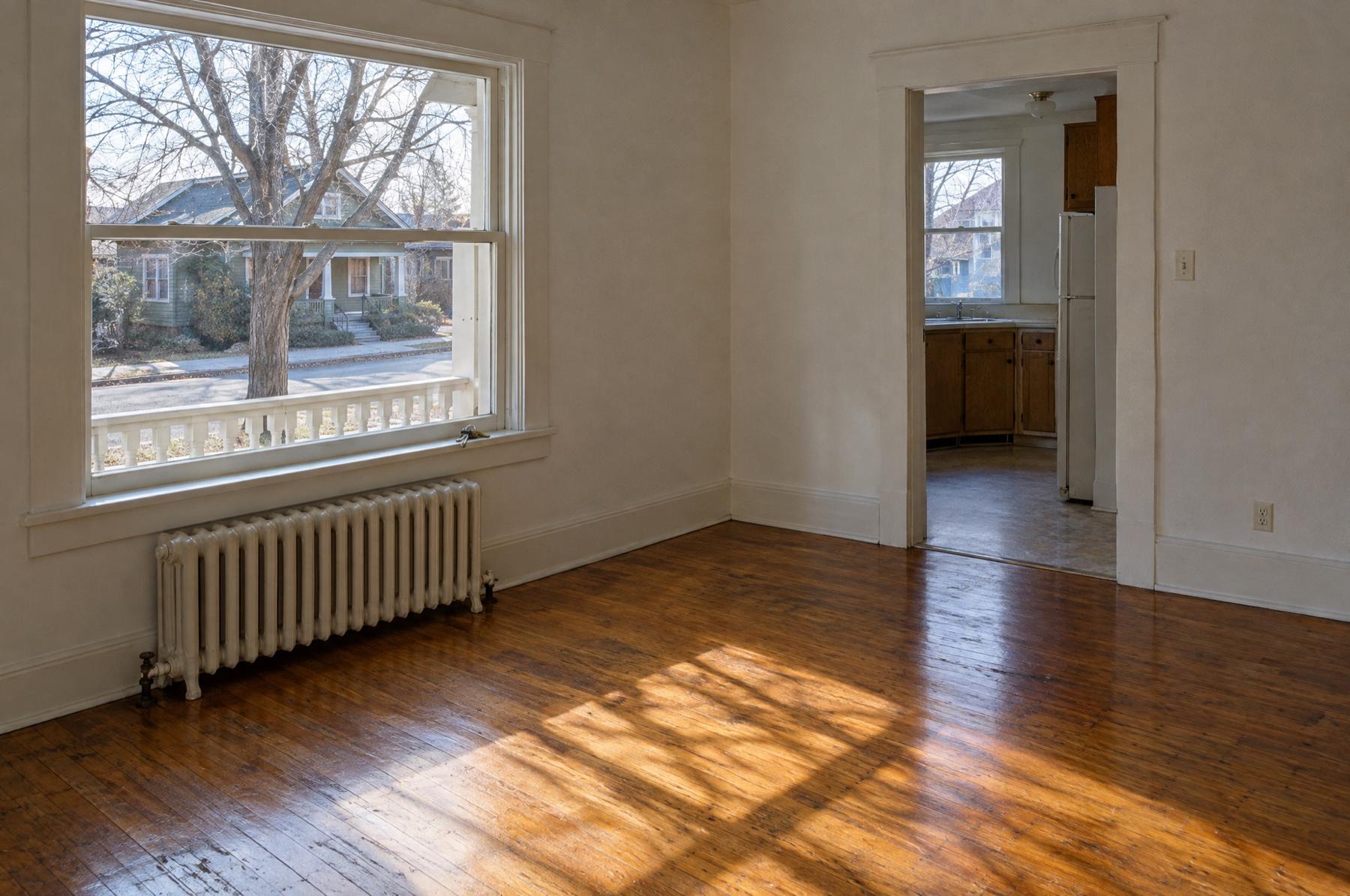 An empty, freshly cleaned living room in an older Spokane rental with oak floors, a radiator under a tall window, and a doorway into the kitchen