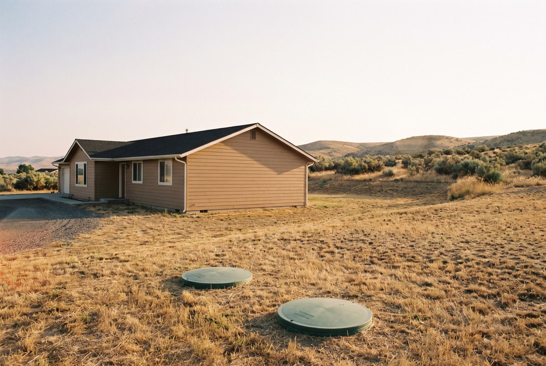 Two green septic tank lids set in a late-summer lawn beside a ranch house on a rural lot in West Richland, with sagebrush hills behind