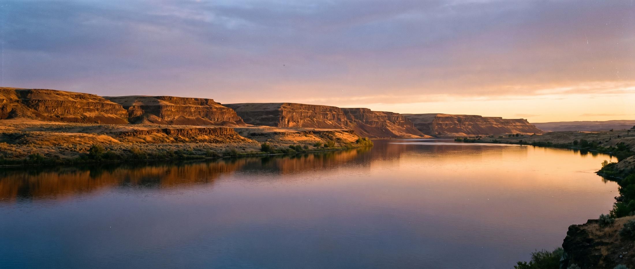 The Columbia River at sunset, calm water reflecting layered basalt bluffs and sagebrush benches along the far shore